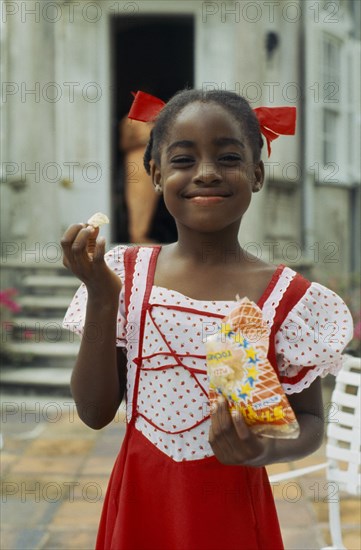 WEST INDIES, Barbados, Easy Hall, Young smiling girl in red and white dress with red ribbons in hair eating crisps from a packet