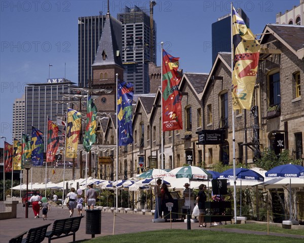 AUSTRALIA, New South Wales, Sydney, The Rocks harbourside area with people walking past restaurants festooned with umbrellas and flags
