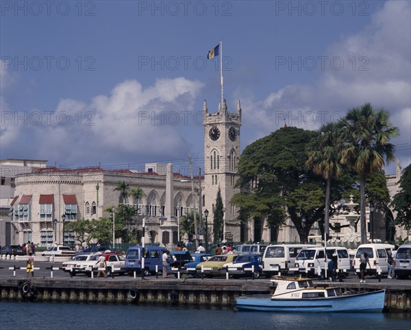 BARBADOS, Bridgetown, The Carenage. Buildings and church clock tower with cars on quay and a boat on water