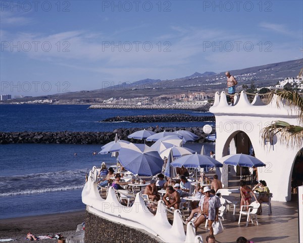 SPAIN, Canary Islands, Tenerife, Playa de las Americas. Terraced bar overlooking the beach with customers sitting at table and a man standing on a balcony above