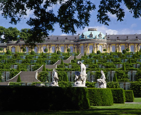 GERMANY, Brandenberg, Potsdam, "Sanssouci Park.  Terraced steps and topiary leading to Palace, with statues in the foreground partly framed by tree branches. "