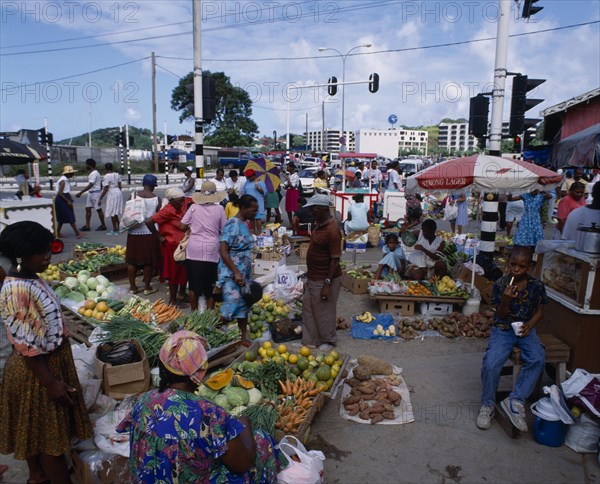 WEST INDIES, St Lucia, Castries, "Fruit and  vegetable street market. Pumpkins, carrots, cabbage, yams, spring onions, bananas, oranges near street with traffic lights and ice-cream vendor in background. "