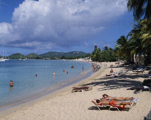 WEST INDIES, St Lucia, Reduit Beach, Sunbathers and swimmers on sandy beach lined with palm trees. Yachts seen on the water and green hills behind