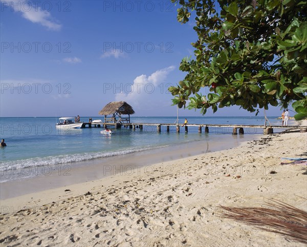 WEST INDIES, Tobago, Pigeon Point, Sandy beach with boats at wooden jetty with tree leaves from right and foot prints in sand