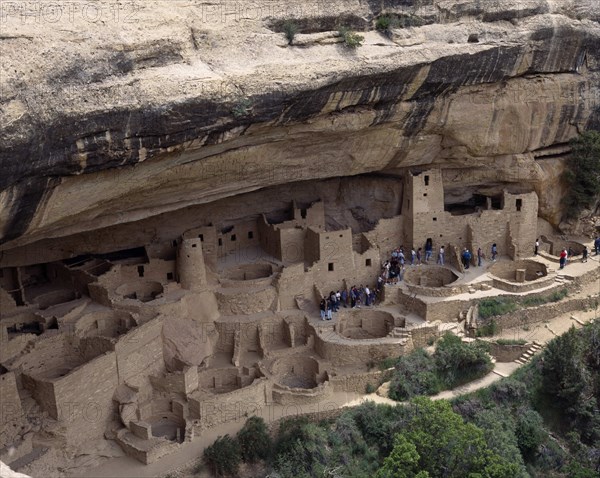 USA, Colorado, Mesa Verde National Park, Cliff Palace the preserved ruins and cave houses beneath rock face. Visitors walk through site of walls and towers with circular foundations leading to deeper caves.