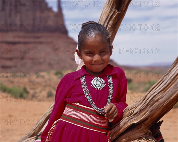 USA, Arizona, Monument Valley, Portrait of Navejo girl wearing a red dress and turquoise necklace and broach leaning against  adead tree trunk.