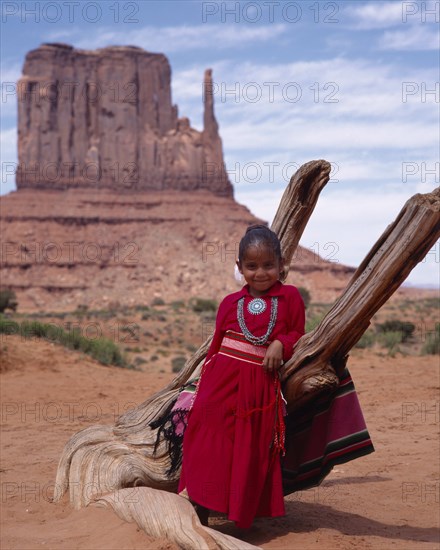 USA, Arizona, Monument Valley, Navejo girl leaning against dead tree wearing a red dress and turquoise necklace and broach with the Mittens rock formation in the background