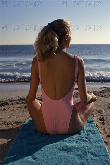 USA, Florida, Sunbathing, Young woman on sandy beach sitting in a meditation position on a green towel looking out towards the sea