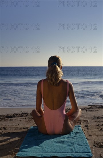 USA, Florida, Sunbathing, Young woman on sandy beach sitting in a meditation position on a green towel looking out towards the sea