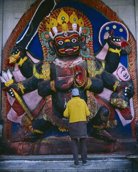 NEPAL, Kathmandu, "Man in front of temple carving of Black Bhairab in Durbar Square depicting Shiva in his most fearsome form with six arms, a head dress of skulls and carrying weapons."