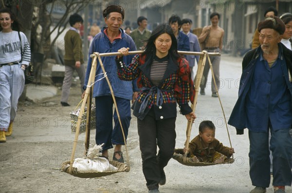CHINA, Guizhou, Bijie, Woman with carrying scales over her shoulders balancing a small child with a sack.