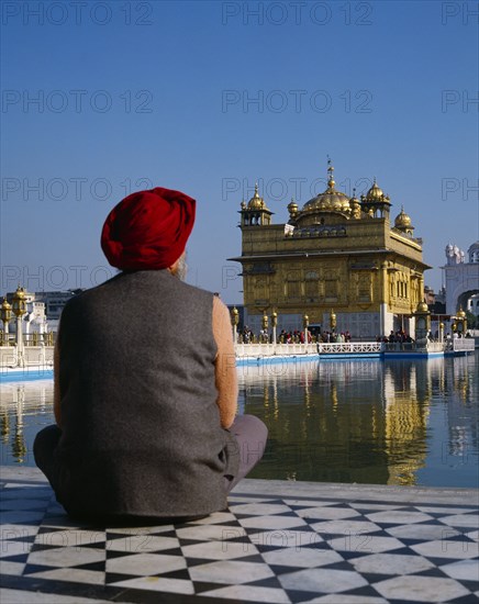 INDIA, Punjab, Amritsar, The Golden Temple.  Man wearing red turban sitting on tiled floor beside lake seen from behind.