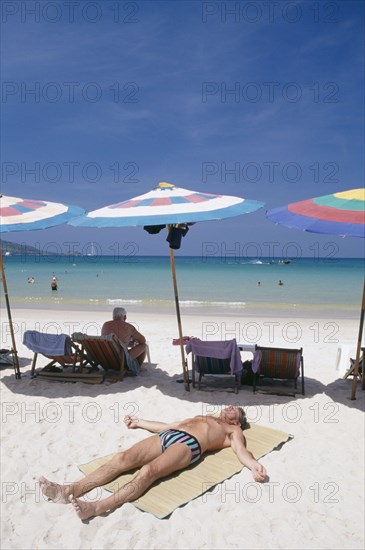 THAILAND, Phuket, Patong Beach, Sun bather lying on the sand in the foreground with row of umbrellas and deck chairs behind and the sea beyond