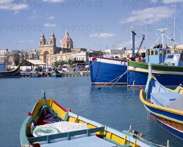 MALTA, Marsaxlokk, The Harbour with the prows of colourful moored fishing boats with the cathedral in the distance