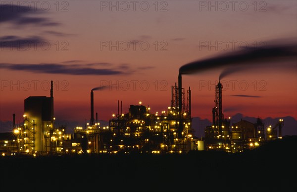 INDUSTRY, Factory, Refinery, Grimsby petrochemical plant illuminated at dusk. Buildings and smoke from chimneys silhouetted against pink and orange sky.
