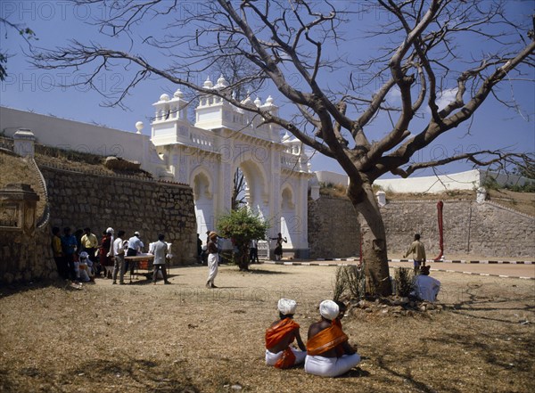 INDIA, Karnataka, Mysore, "Maharaja’s Palace.  Exterior wall and white, carved and decorated entrance arch with people gathered in dry, grassy area in foreground."