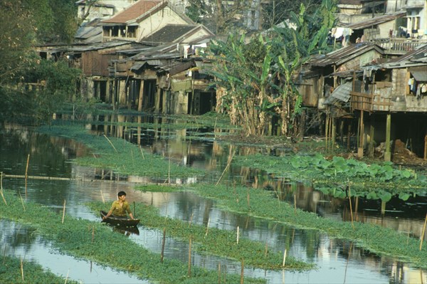 VIETNAM, Central, Hue, Wooden houses raised on stilts above waterway with man in small canoe in foreground.