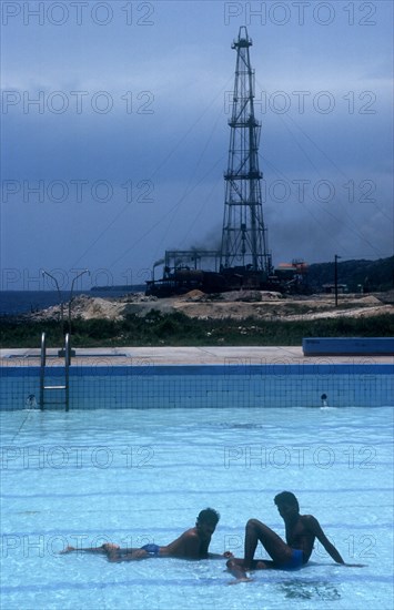 CUBA, Tibacoa Resort , Two men sitting in a shallow swimming pool with Oil derrick in the background