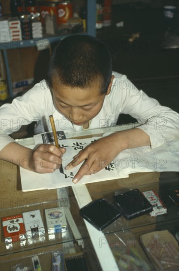 CHINA, Ningxia Province, Writing, Boy practising writing Chinese characters in shop owned and run by his parents.