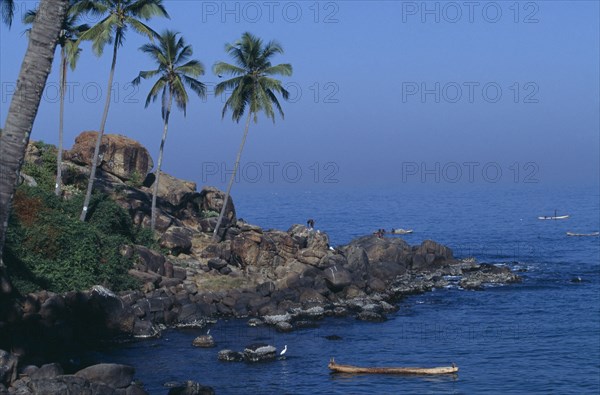INDIA, Kerala, Kovalam, Coastal scene with coconut palms on rocky promontory and men in boats beyond
