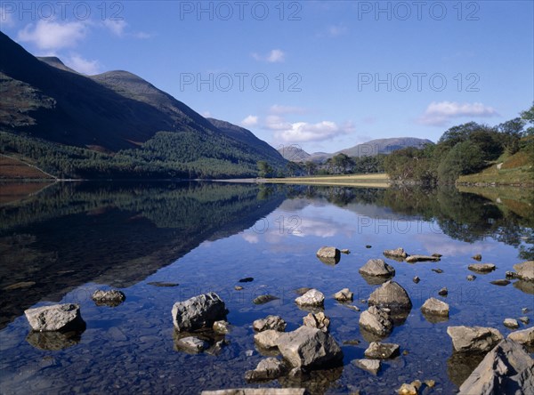 ENGLAND, Cumbria, Lake District, "Buttermere.  Landscape, blue sky and drifting cloud reflected in lake with small rocks protruding from clear, shallow water in the foreground."