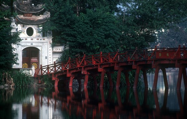 VIETNAM, Hanoi, Hoan Kiem Lake. View of red painted bridge across water to decorative building surrounded by lush vegetation.