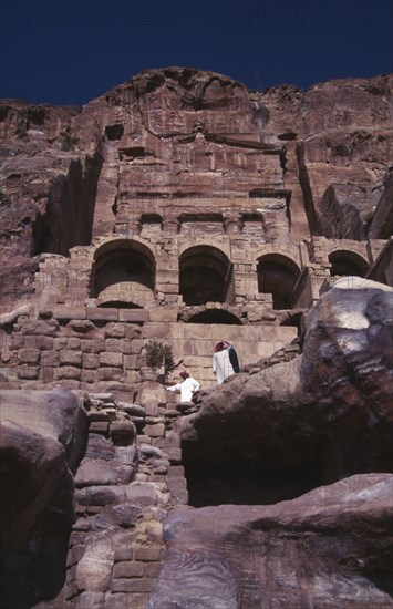 JORDAN, Petra, View looking up at historical tombs carved in sandstone cliffs with two men dressed in white looking downwards