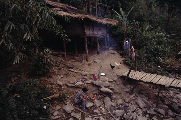 THAILAND, North, Mae Sariang, Karen refugee camp house with chickens outside by wooden bridge and boy tending a fire in the foreground