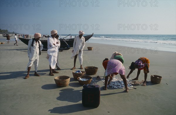 INDIA, Goa, Colva , Three men in turbans watch three women sorting the morning catch near a boat on the beach.