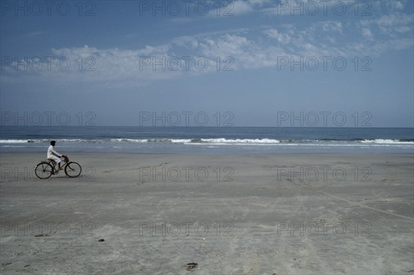 INDIA, Goa, Colva Beach , Cyclist riding along sand of the empty beach with waves rolling on to the shore