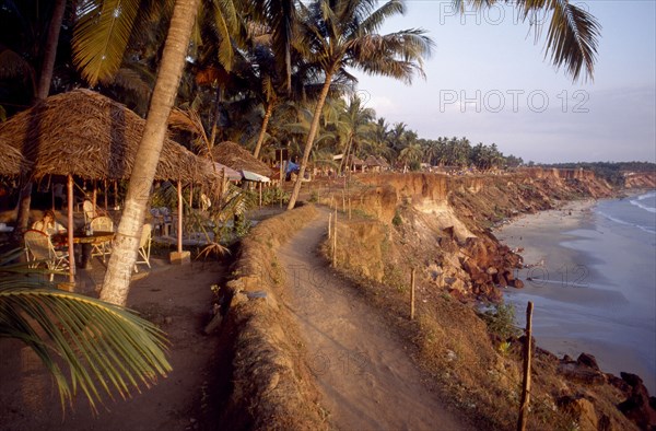 INDIA, Kerala, Varkala , Coastal path along cliff tops with sandy beach below and woman sitting under thatched restaurant shade