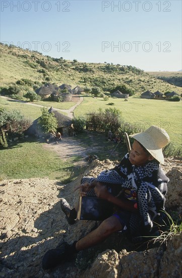 SOUTH AFRICA, Qwa-Qwa, Basotho Village, "Basotho Cultural Village, local man playing an instument called a Sekoankula, hills and huts in background. "