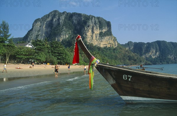 THAILAND, Krabi, Ao Nang Beach, Long tail boat moored in shallow water at the edge of beach with tourists.  Forest covered cliffs behind.