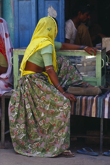 INDIA, Rajasthan, Devgarh , "Woman in rose patterned sari and yellow head covering wearing ankle and toe ringsand bracelets, sitting outside jewellery shop. "
