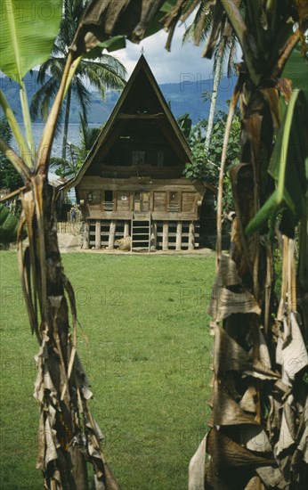 INDONESIA, Sumatra, Lake Toba , Wooden Batak house with pointed roof on Samosir Island seen through banana trees