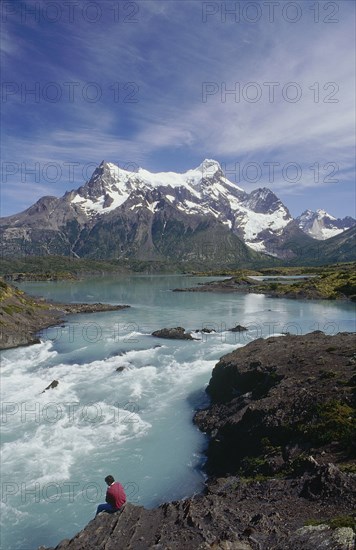 CHILE, Patagonia, Torres Del Paine, Man sitting on the riverbank beside rapids below the distant snow covered mountains