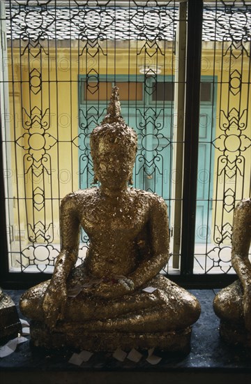 THAILAND, Bangkok, Seated Buddha covered with gold leaf in front of window with decorative metal shutter.