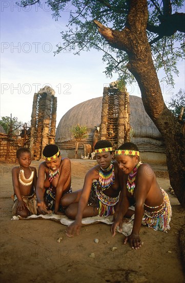 SOUTH AFRICA, KwaZulu Natal, Shakaland , Local children in traditional dress playing a game with stones on the ground outside thatched huts