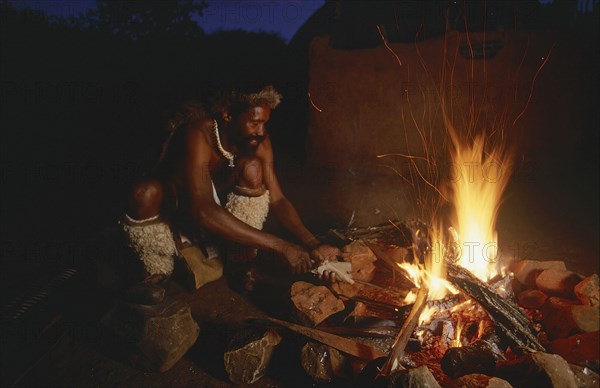 SOUTH AFRICA, Kwa Zulu Natal , Eshowe, Zulu man making a spear over an open fire at night outside thatched huts