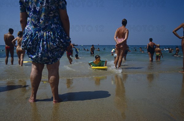 FRANCE, Aquitaine, Pyrenees Atlantiques, Biarritz.  Busy beach scene with people standing and playing in the water. Boy on yellow board and lady wearing a dress.