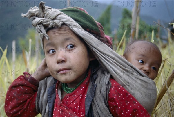 NEPAL, Bung, Rai girl carrying baby in sling on her back supported by cloth knotted around head.