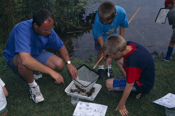 EDUCATION , Primary, Field Trip, "Pond Dipping activity. Children with adult inspecting a fishing net to identify species of aquatic life from worksheets in ecological study . Norfolk, England."