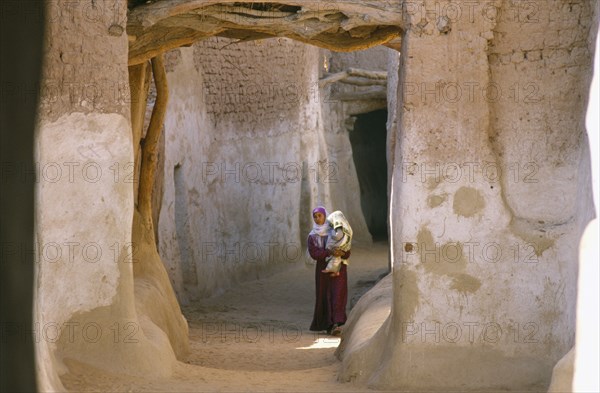 EGYPT, Western Desert, Dhalla Oasis, Woman carrying child along street between mud brick buildings.