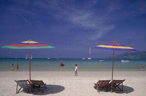 THAILAND, Phuket, Patong Beach, Empty deckchairs and sun parasols on the beach with people walking by the water