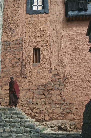 TIBET, Xigatse, Tashilumbo Monastery, Monk standing on steps in front of peach coloured painted wall