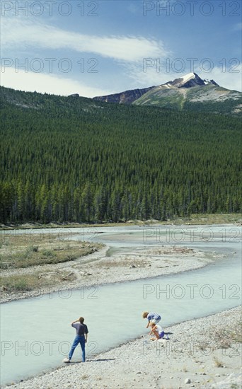 CANADA, British Columbia, Jasper National Park, Visitors beside river looking towards dense pine forest with jagged mountain peak above.