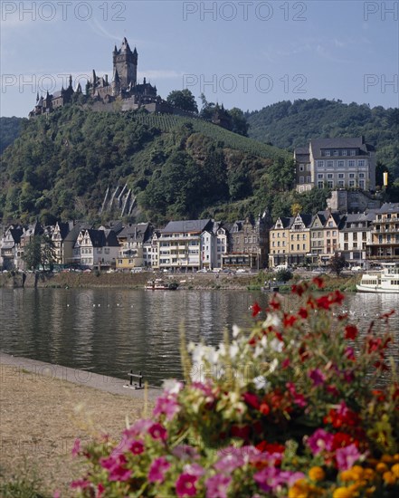 GERMANY, Rhineland, Cochem, Hilltop castle overlooking houses beside the Mosel River with flowers in the foreground