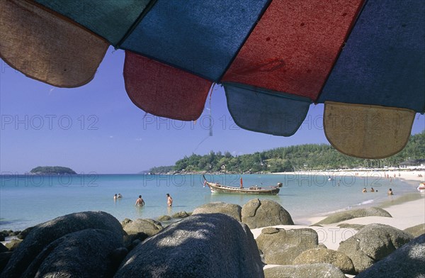 THAILAND, Phuket, Kata Noi Beach, "Little Kata Beach seen from beneath a colourful umbrella with rocks, a boat and people wading in the sea "