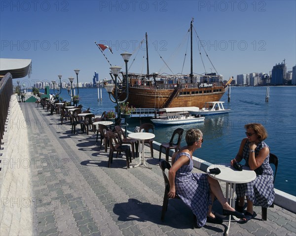 UAE, Abu Dhabi, Corniche. Restaurant Dhow by seam moored boats with two woman sat on table and chairs in the foreground