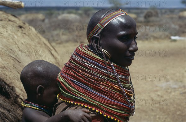 KENYA, North Frontier, Baragoi, Samburu woman wearing traditional multi-strand necklaces with child on her back.